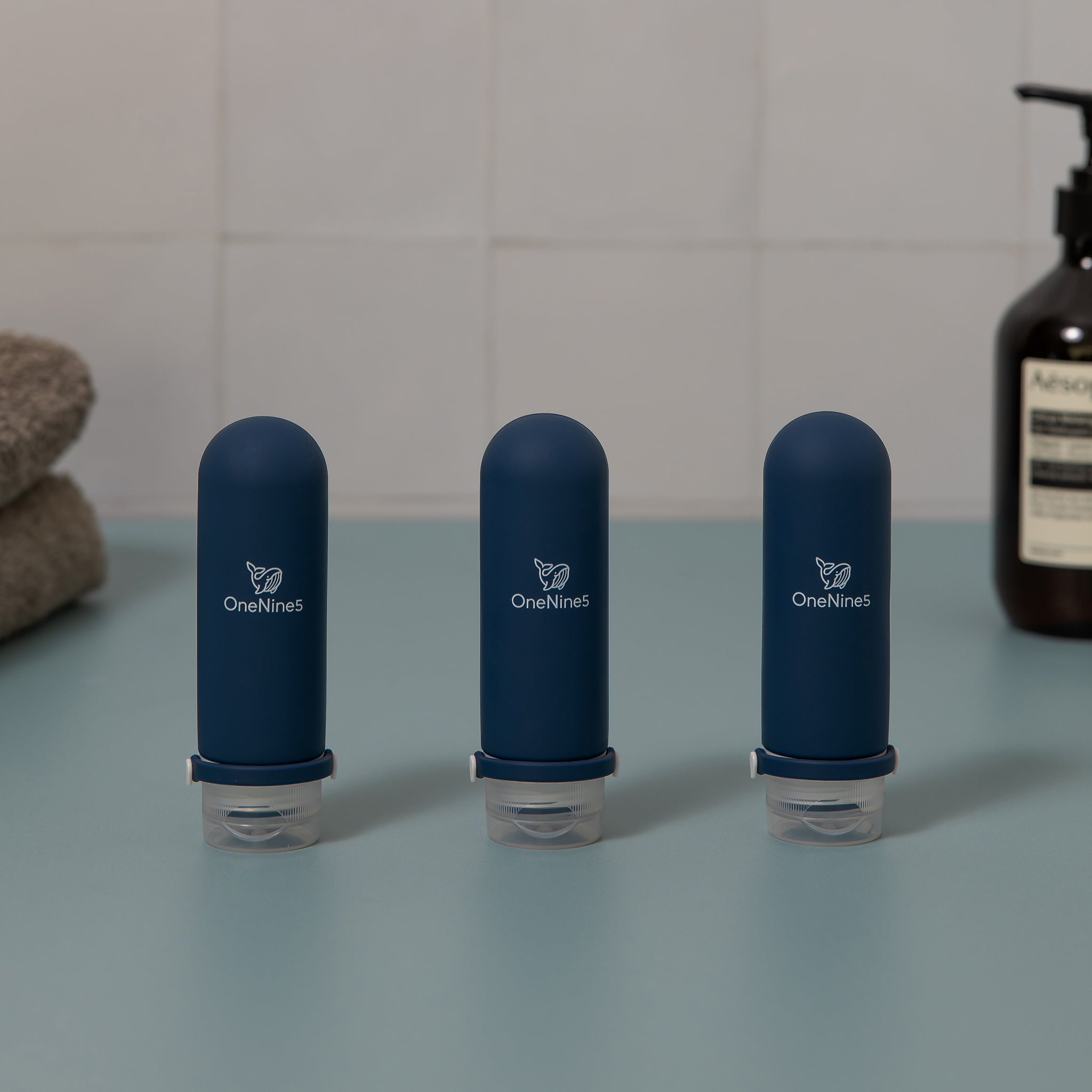 Three blue OneNine5 silicone travel bottles on green and white tiled background. Blurred out in the background is a towel and bottle of body wash. A white OneNine5 logo is visible on the front of the bottles.