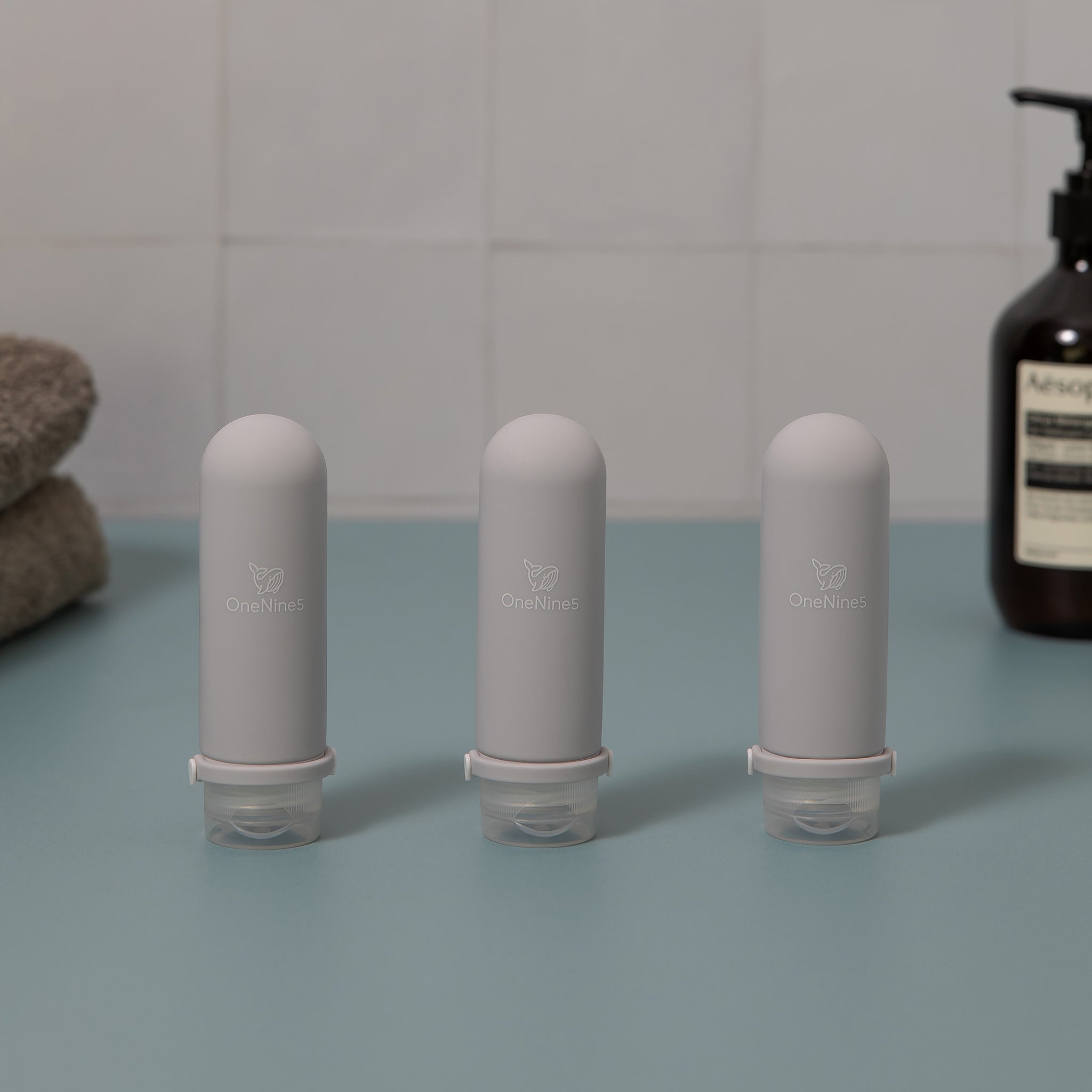 Three grey OneNine5 silicone travel bottles on green and white tiled background. Blurred out in the background is a towel and bottle of body wash. A white OneNine5 logo is visible on the front of the bottles.