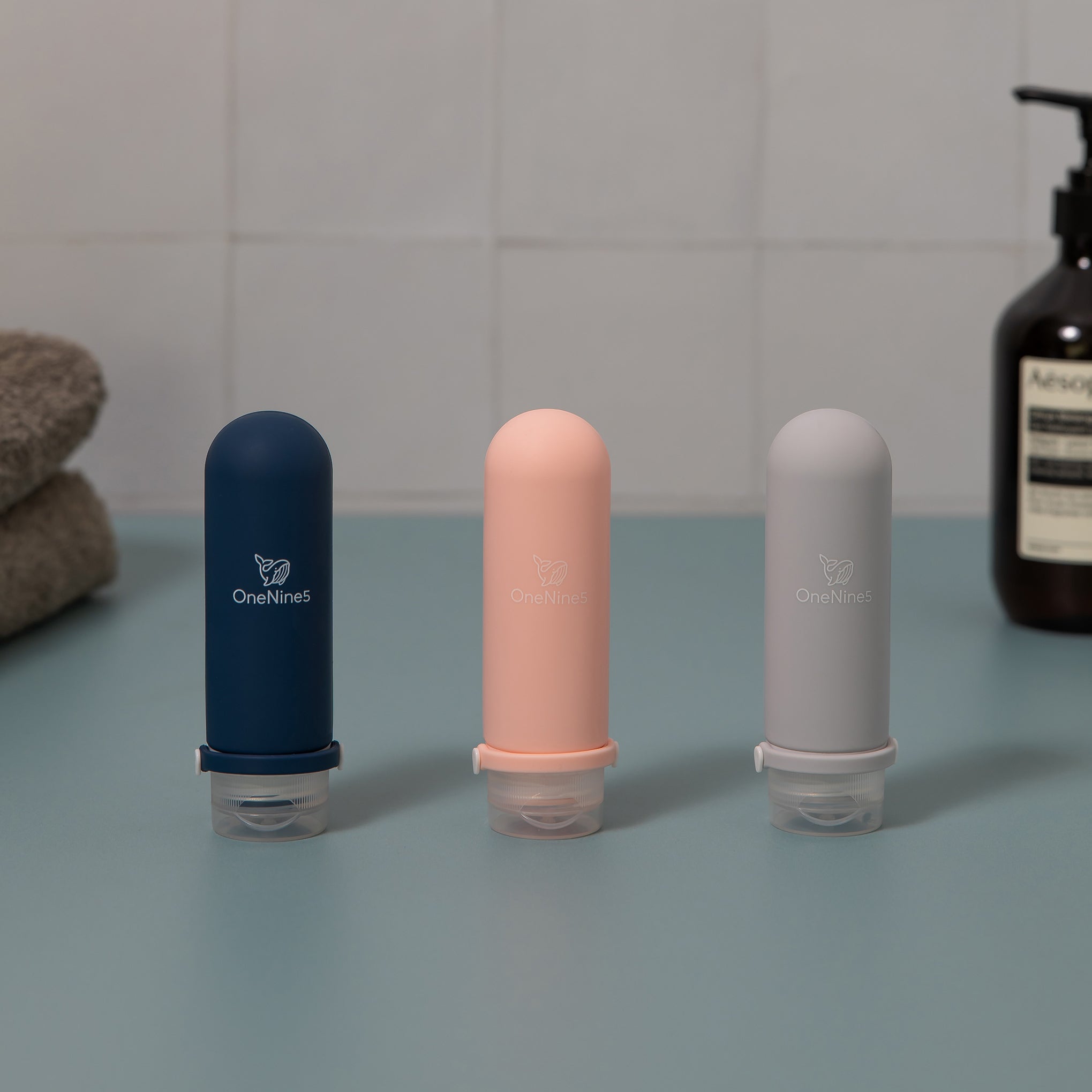 A blue, pink & grey OneNine5 silicone travel bottle on green and white tiled background. Blurred out in the background is a towel and bottle of body wash. A white OneNine5 logo is visible on the front of the bottles.