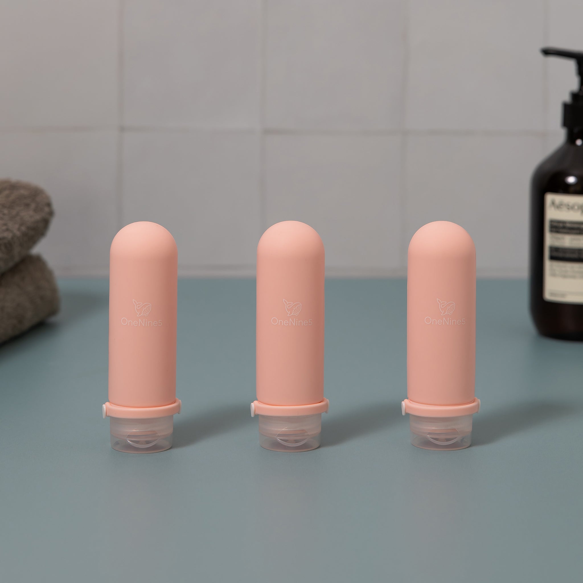 Three pink OneNine5 silicone travel bottles on green and white tiled background. Blurred out in the background is a towel and bottle of body wash. A white OneNine5 logo is visible on the front of the bottles.