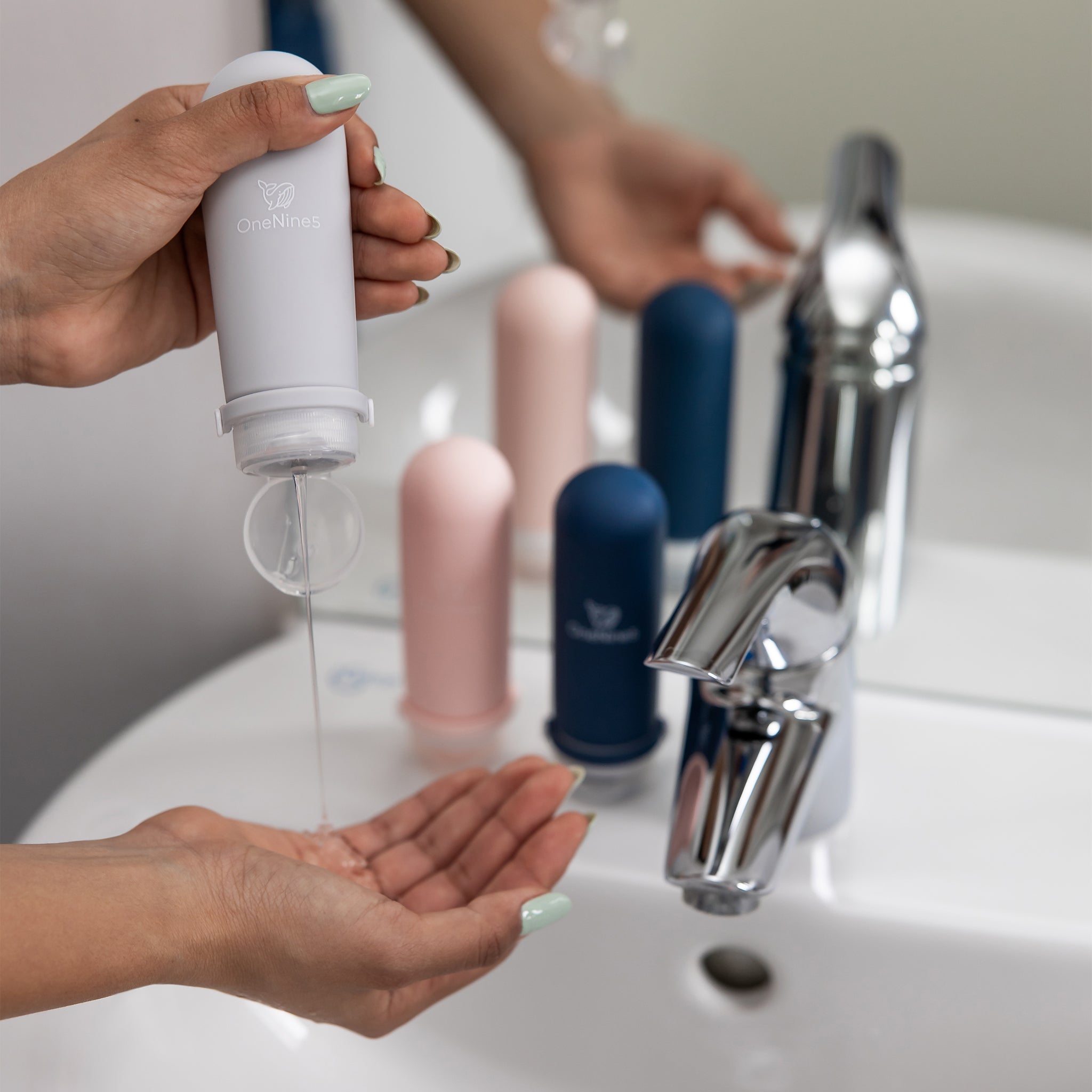 A female in a bathroom, squeezing the soft silicone OneNine5 grey travel bottle into her hand, over the sink. With the leakproof cap (lid) flipped open. Blurred in the background is a blue and pink silicone bottle resting on the sink.