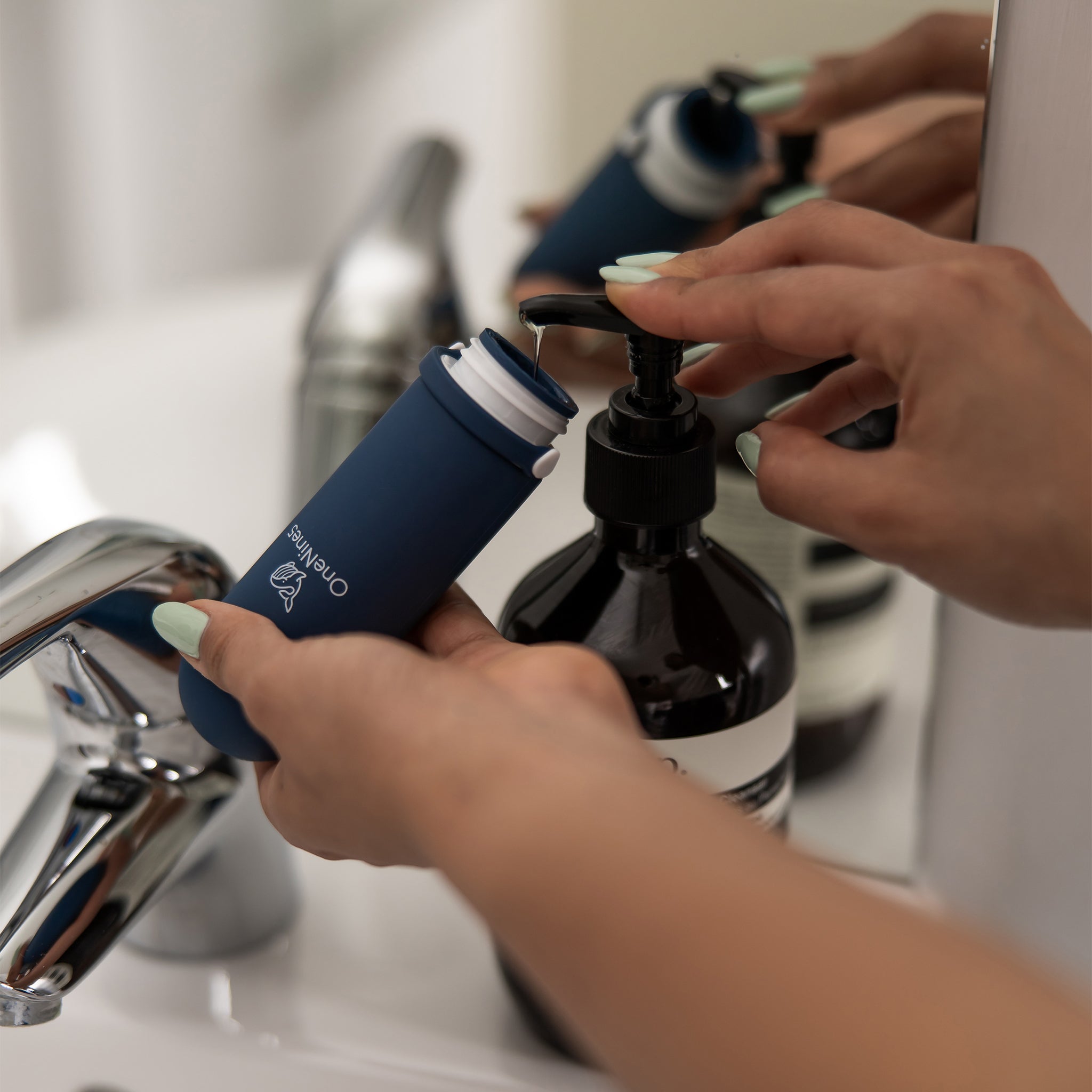 A female in a bathroom, pumping Aesop body wash into the blue OneNine5 silicone bottle. The flip cap is removed to ensure easy refill into the wide neck.