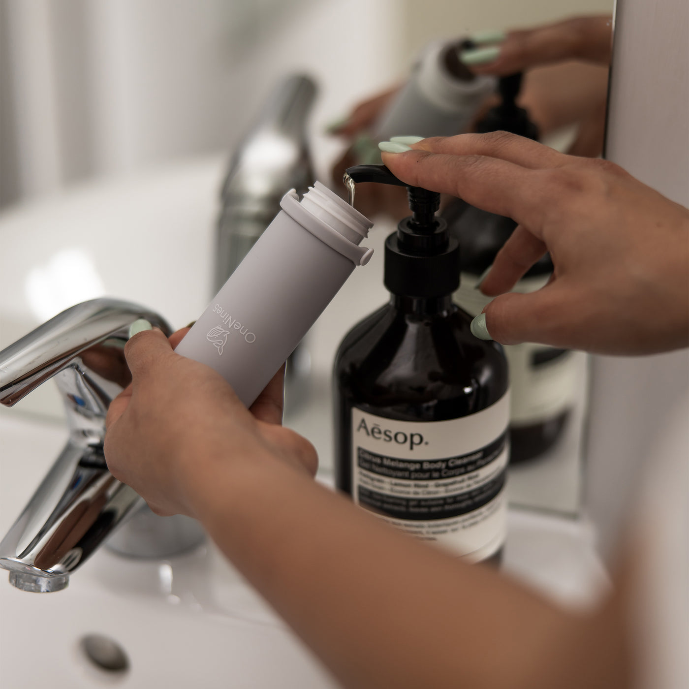 A female in a bathroom, refilling the grey OneNine5 silicone travel bottle with Aesop body wash, from a pump bottle.