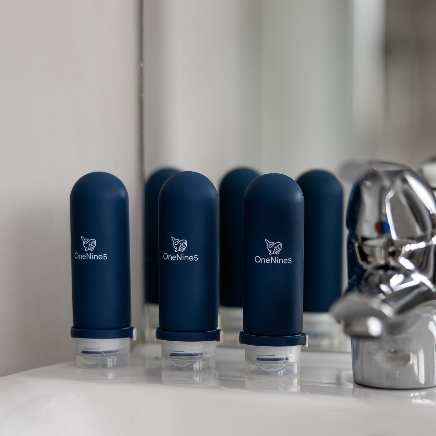 -pack of blue silicone travel bottles on a white bathroom sink next to a chrome tap.