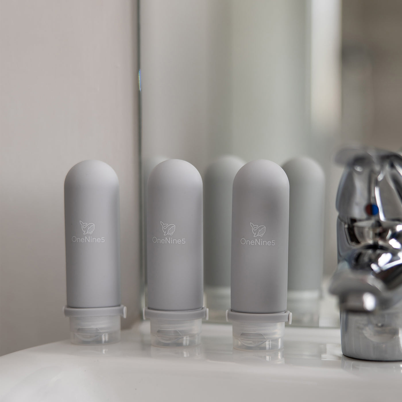 3-pack of grey silicone travel bottles on a white bathroom sink next to a chrome tap.