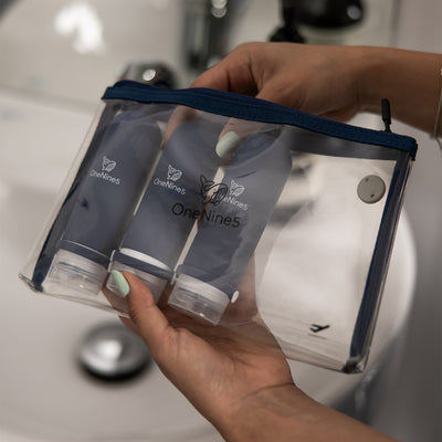 A woman, in the bathroom, packing 3 blue silicone bottles inside the removable, clear OneNine5 toiletry/ cosmetic bag.