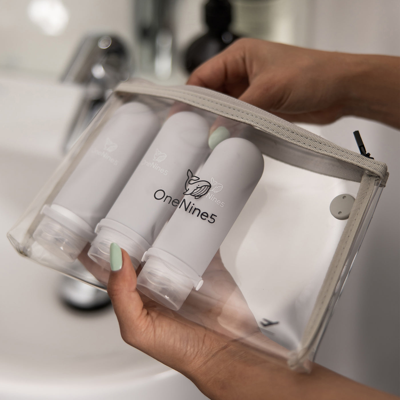 A woman, in the bathroom, packing 3 grey silicone bottles inside the removable, clear OneNine5 toiletry/ cosmetic bag.