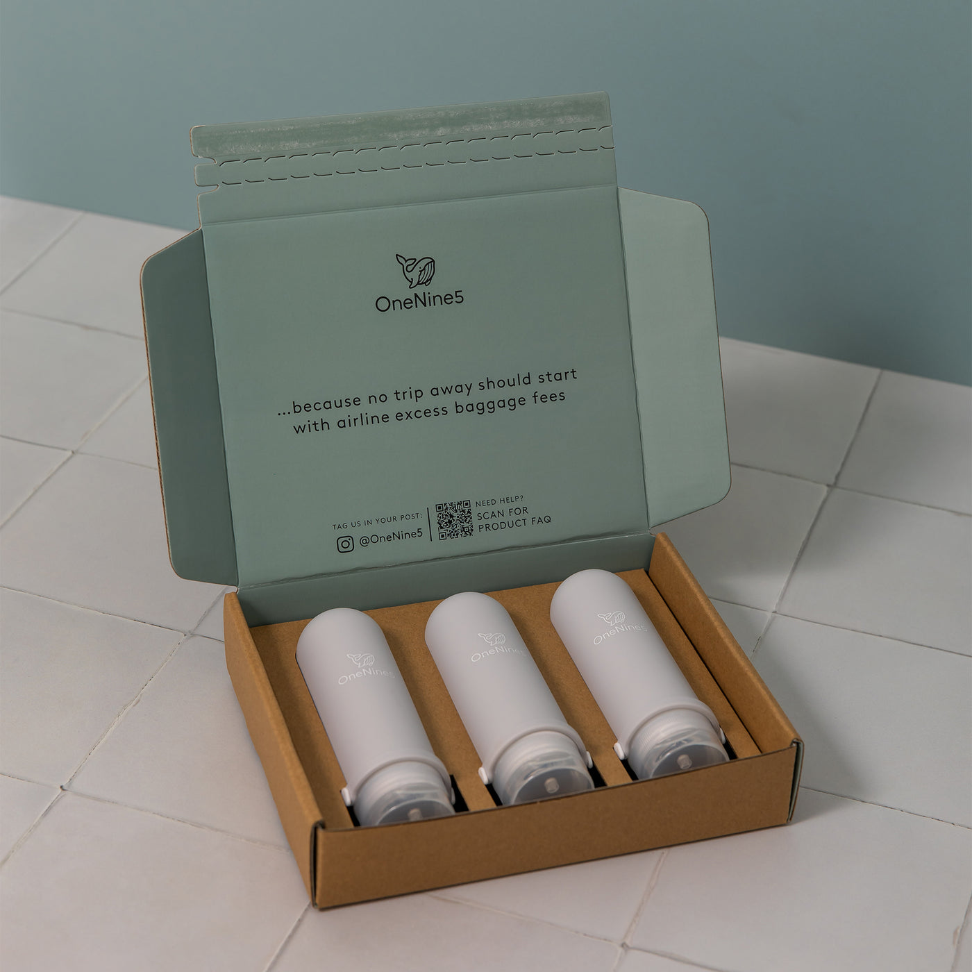 3 pack of grey silicone travel bottles are packed inside a brown, recyclable kraft paper box. Printed inside the packaging is the OneNine5 logo, a QR code and the caption ‘Because no trip away should start with airline excess baggage fees’.