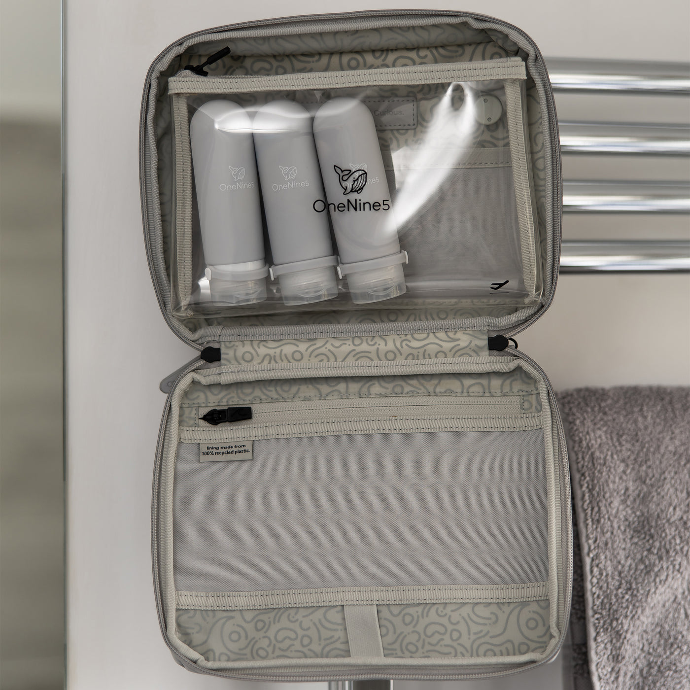 Three grey silicone travel bottles inside the clear toiletry pouch. The pouch is attached inside the OneNine5, Moeraki Grey wash bag, using the hanging hook, in the bathroom.