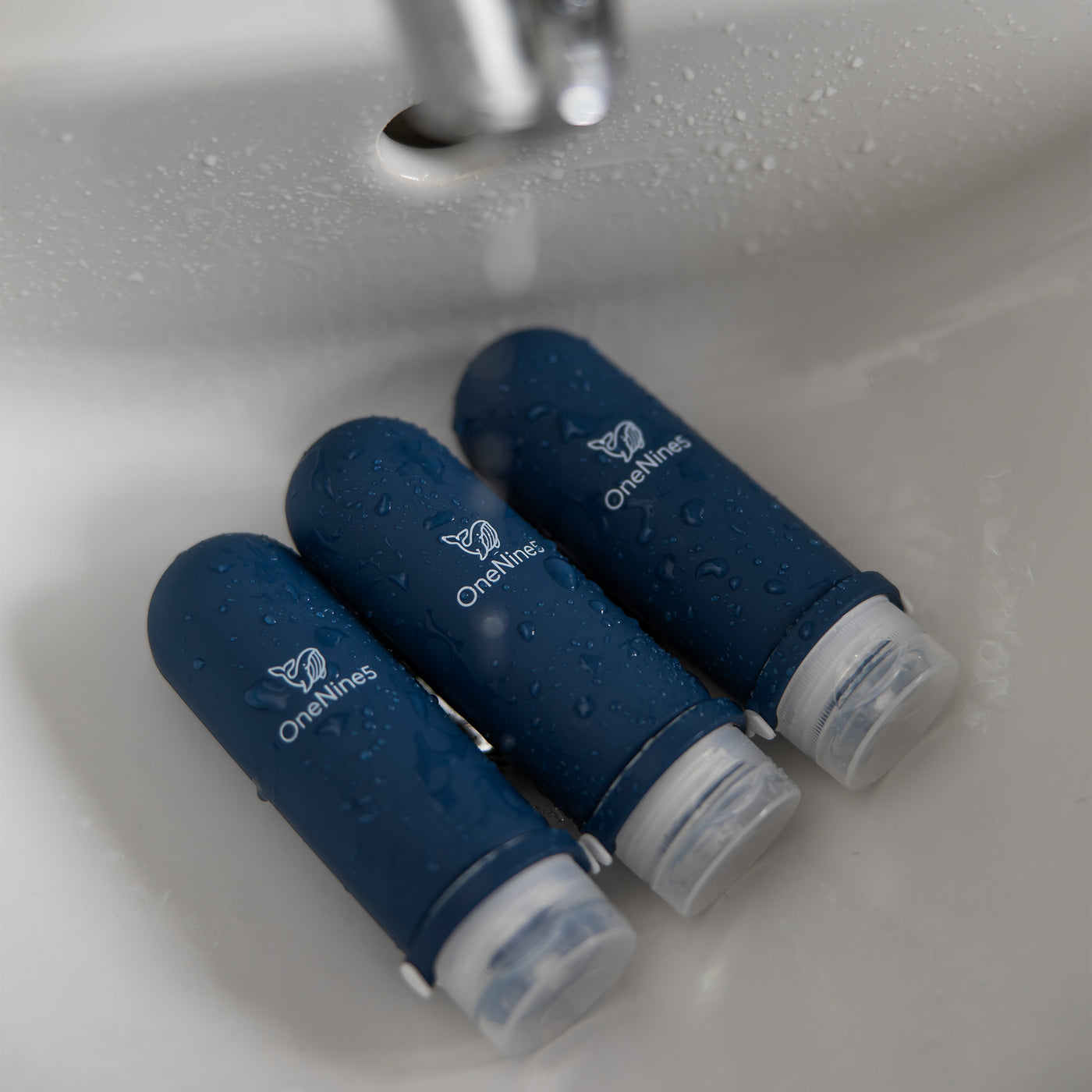 Three blue silicone travel bottles in the bathroom sink. A white OneNine5 logo is visible on the reusable bottles and they are being splashed with water.