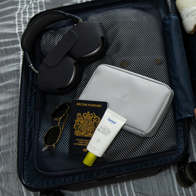 A Moeraki Grey Eco Essentials Pouch being packed on an open suitcase, on a bed. Next to Apple Airpods Max, a UK passport, Supergoop SFD and Ray Ban sunglasses