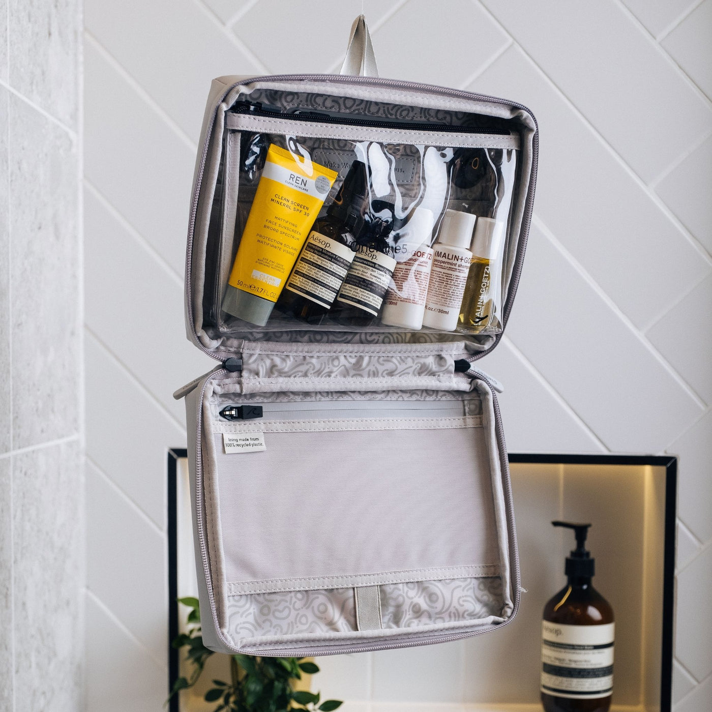 A Moeraki Grey OneNine5 Wash Bag zipped open and hanging up in the bathroom. The wash bag is packed with a range of toiletries and cosmetics