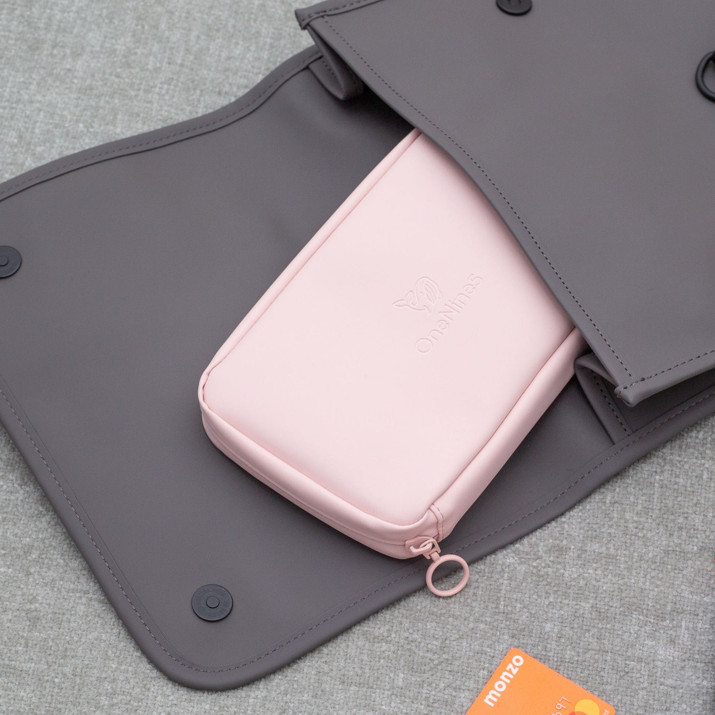 A Komodo Pink OneNine5 Essentials Pouch poking out the top of a backpack, on the bed.